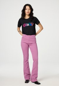 Zwarte t-shirt met kleurrijk, textuurvol "CHAOT" ontwerp; roze flare jeans; model staat met handen op de heupen tegen een witte achtergrond.