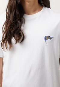 Hvid bomulds t-shirt med korte ærmer, med et broderet flag-design i blå og gul og teksten "CHUCK TAYLOR" på venstre bryst.