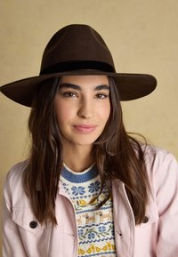Femme aux longs cheveux bruns portant un chapeau marron à large bord, un pull à motifs bleus et jaunes, et une veste rose clair boutonnée.