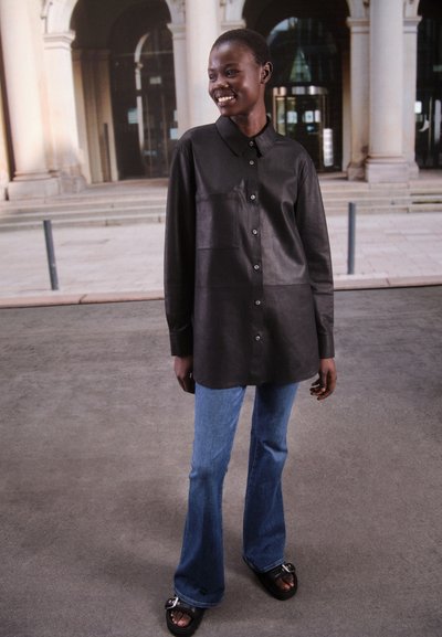 Camicia nera in pelle con chiusura a bottoni, design con colletto e tasca frontale, abbinata a jeans in denim blu svasati e sandali neri.