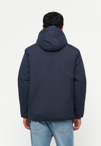 Man wearing a navy blue hooded jacket and light blue jeans, shown from behind against a plain white background.
