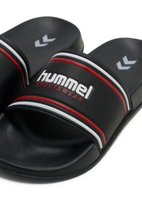 Black slide sandals with red and white stripes, featuring a textured strap and "hummel SPORTSWEAR" logo on the band. Flat sole design.
