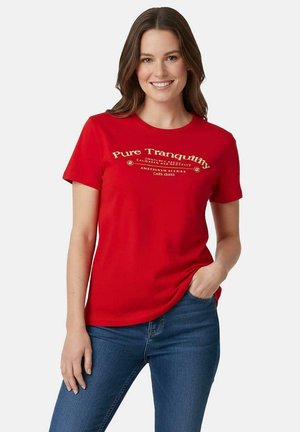 Vrolijk glimlachende vrouw met een rood "Pure Tranquility" T-shirt en blauwe spijkerbroek, staand met één hand in haar zak tegen een effen achtergrond.