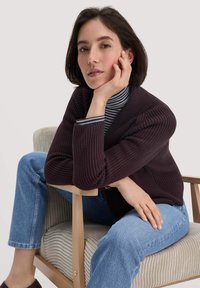 Dunkelpurpurfarbener Strickcardigan mit geripptem Muster. Kombiniert mit einem gestreiften Rollkragenpullover und hellblauen Jeans. Person sitzt auf einem Holzstuhl.