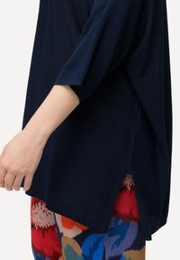 Dunkelblaues, kurzärmliges Oberteil mit lockerer Passform und seitlichen Schlitzen. Die Textur wirkt gerippt. Kombiniert mit bunten gemusterten Hosen.