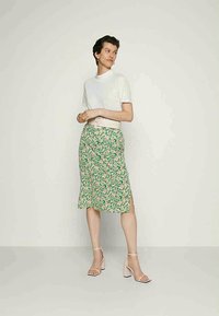 Mulher vestindo uma blusa branca de mangas curtas, saia midi floral verde com fenda lateral, cinto largo branco e sandálias de salto bloco nuas, encostada a uma parede lisa.