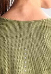 Locker sitzendes, olivgrünes Oberteil aus strukturiertem Stoff mit weitem Ausschnitt und fünf dekorativen weißen Punkten entlang des Rückens.