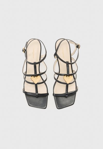 Sandalias de cuero negro con punta cuadrada, múltiples correas y un acento en forma de corazón dorado en el centro. Incluye una correa ajustable para el tobillo.