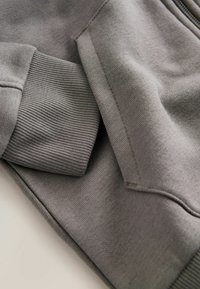 Grauer Sweatshirtstoff mit glatter Textur, ausgestattet mit gerippten Bündchen und einer Seitentasche. Nahaufnahme von Nähten und Designdetails.