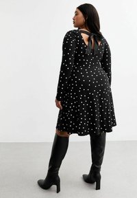 Robe noire à manches longues à pois blancs, avec un détail de nouage dans le dos, jupe évasée, portée avec des bottes noires montantes.