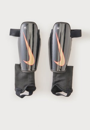 Paar schwarze Schienbeinschoner mit orangefarbenen Nike-Swoosh-Logos und befestigten schwarzen Knöchelärmeln auf hellem Hintergrund.