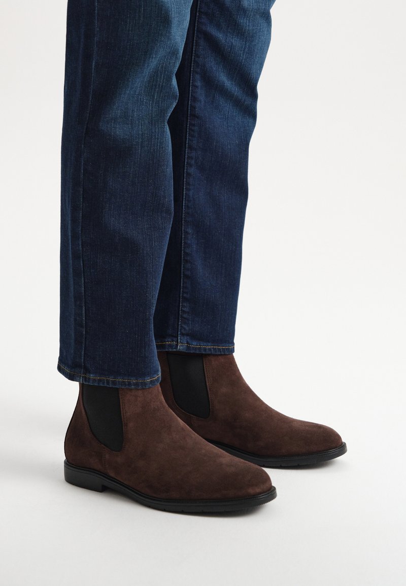 Bruna mockaskinnsankelboots med elastiska sidopaneler, rundade tår och en låg svart gummihäl, bärs med mörka denimjeans.