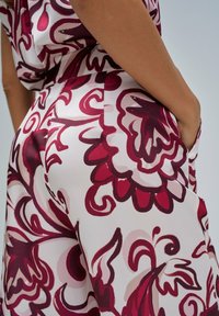 Mujer vestida con pantalones y blusa blanca con grandes patrones florales rojos y rosados, con una mano en el bolsillo, fondo neutral.
