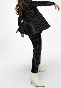 Černý oblek s pinstripem zahrnující sako a kalhoty; sako má rozšířený design, bílé pruhy a hladkou texturu. V kombinaci se smetanovými botami s špičatou špičkou.