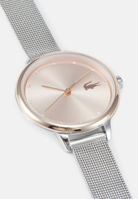 Reloj plateado redondo con esfera metálica cepillada, acentos en oro rosa y correa de malla. Presenta pequeños cristales que marcan las posiciones de las horas.