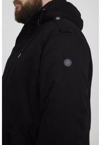 Svart hettejakke med en glatt tekstur, med trykknapper, justerbare snorer og et sirkulært logo-patch på venstre skulder.