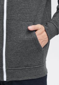 Sudadera gris con cremallera hecha de una tela suave, con una cremallera blanca, bolsillos delanteros y puños acanalados. La textura parece lisa y casual.