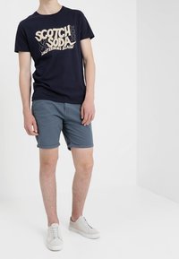 Marineblauwe T-shirt met lichtbeige grafisch ontwerp; grijze korte broek; witte sneakers. De outfit toont een casual, ontspannen pasvorm en moderne stijl.