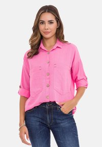 Rosa Leinenhemd mit Knopfverschluss, zwei Fronttaschen, hochgerollten Ärmeln und beigen Knöpfen. Kombiniert mit dunkelblauen Jeans.
