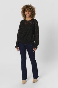 Schwarzes Spitzentop mit langen Ärmeln und Rüschensaum, das überaus feine florale Ausschnitte aufweist. Kombiniert mit dunkelblauen Schlagjeans und hellbraunen Schuhen.