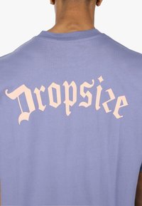 Lilla bomull T-skjorte med stor, stilisert beige tekst "Dropsize" trykt på baksiden. Klassisk rund hals og korte ermer.