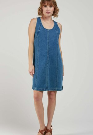 Femme aux cheveux courts et ondulés, portant une robe en jean bleu sans manches et des sandales marron, debout devant un fond clair uni.