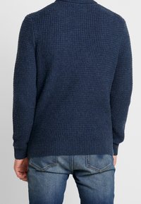 Marinblå texturerad tröja med ribbad design, hög krage och långa ärmar, sedd bakifrån, kombinerad med denimjeans.