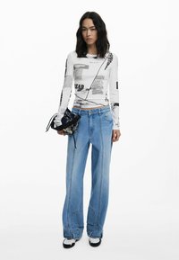 Langärmliges weißes Oberteil mit grafischen Drucken, kombiniert mit weit geschnittenen, hellblauen Jeans. Die Accessoires umfassen eine kleine schwarze Tasche mit aufwendigen Details.