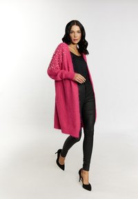 Cardigan rosa esponjoso con acentos de perlas, sobre una blusa negra y pantalones de cuero sintético, combinado con zapatos de tacón negros, en un fondo neutro.