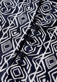 Textil de patrones en blanco y negro que presenta formas geométricas y líneas, con botones oscuros a lo largo de una prenda con cuello. Textura suave y lisa.
