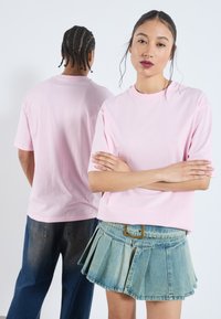 Lys rosa bomulls T-skjorte med korte ermer, vist på en modell. En annen person i mørke jeans vises i bakgrunnen.