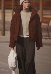 Giacca imbottita marrone, dolcevita a coste grigio chiaro, pantaloni neri e borsa bianca pelosa. Modello con berretto lavorato a maglia grigio, cammina all'aperto.