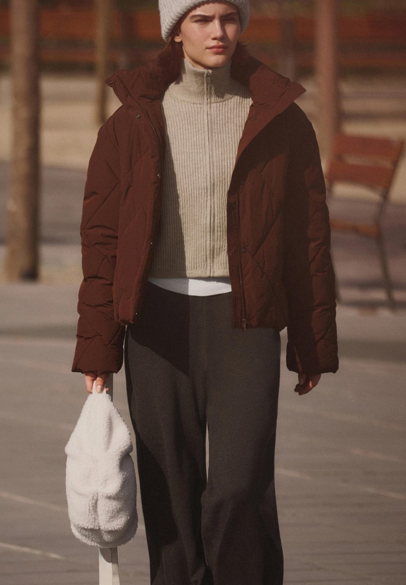 Giacca imbottita marrone, dolcevita a coste grigio chiaro, pantaloni neri e borsa bianca pelosa. Modello con berretto lavorato a maglia grigio, cammina all'aperto.