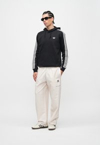 Μαύρο φούτερ με κουκούλα και λευκές ρίγες, μπεζ παντελόνι φόρμας με λογότυπο και μαύρα γυαλιά ηλίου. Τα παπούτσια είναι λευκά με μαύρες λεπτομέρειες.