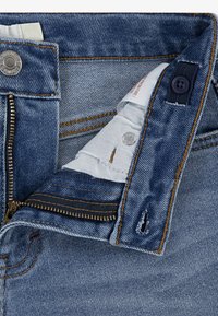 Denim jeans with a zipper fly, copper rivets, stitched seams, and a light blue wash. Interior fabric visible, featuring contrasting color details.