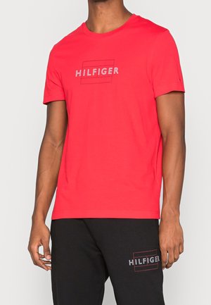 Camiseta roja de algodón de manga corta con un diseño de logotipo en contorno blanco "HILFIGER" en el frente. Combinada con pantalones negros que exhiben el mismo logotipo.