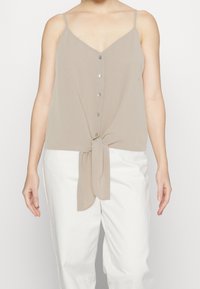 Mujer con top sin mangas beige abotonado con lazo frontal y pantalones blancos, mostrada desde los hombros hasta las caderas contra un fondo liso.