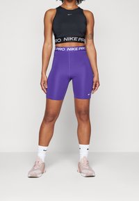 Crop top de sport noir Nike avec bande logo, short taille haute violet, chaussettes blanches et baskets rose clair. Tissu lisse et ajusté.