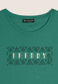 T-shirt verde con scollatura rotonda e una stampa decorativa e testurizzata della parola "FREDDY" in oro metallico su uno sfondo decorato.