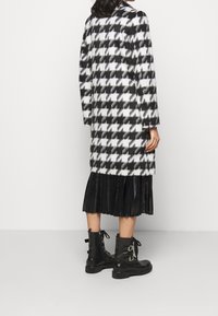Sort-hvid hundetandsmønstret uldfrakke med lange ærmer, knælængde, kombineret med en sort plisseret nederdel og sorte ankelstøvler.