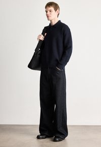 Pulôver de malha canelada azul-marinho com colarinho polo, combinado com calças de ganga escuras de perna larga. A modelo segura uma mala de ombro preta. Texturas suaves.