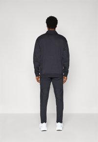 Schwarzes Sweatshirt- und Hosen-Set, das eine lockere Passform aufweist, strukturierte Akzente an der Hose hat und mit weißen Turnschuhen kombiniert ist.