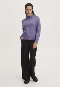 Lilla strikket sweater med høj hals, parret med sorte brede bukser. Stoffet ser blødt og let tekstureret ud.