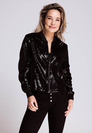 Schwarze Bomberjacke mit Reißverschluss, Paillettenstruktur und gerippten Bündchen, kombiniert mit schwarzen Hosen mit silbernen Knopfdetails.