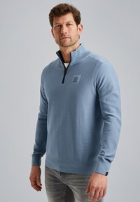  Lichtblauwe kwart-rits pullover met een gestructureerde afwerking, geribde zoom en manchetten, en een klein logo-opdruk op de borst. Gemaakt met een paar grijze jeans.