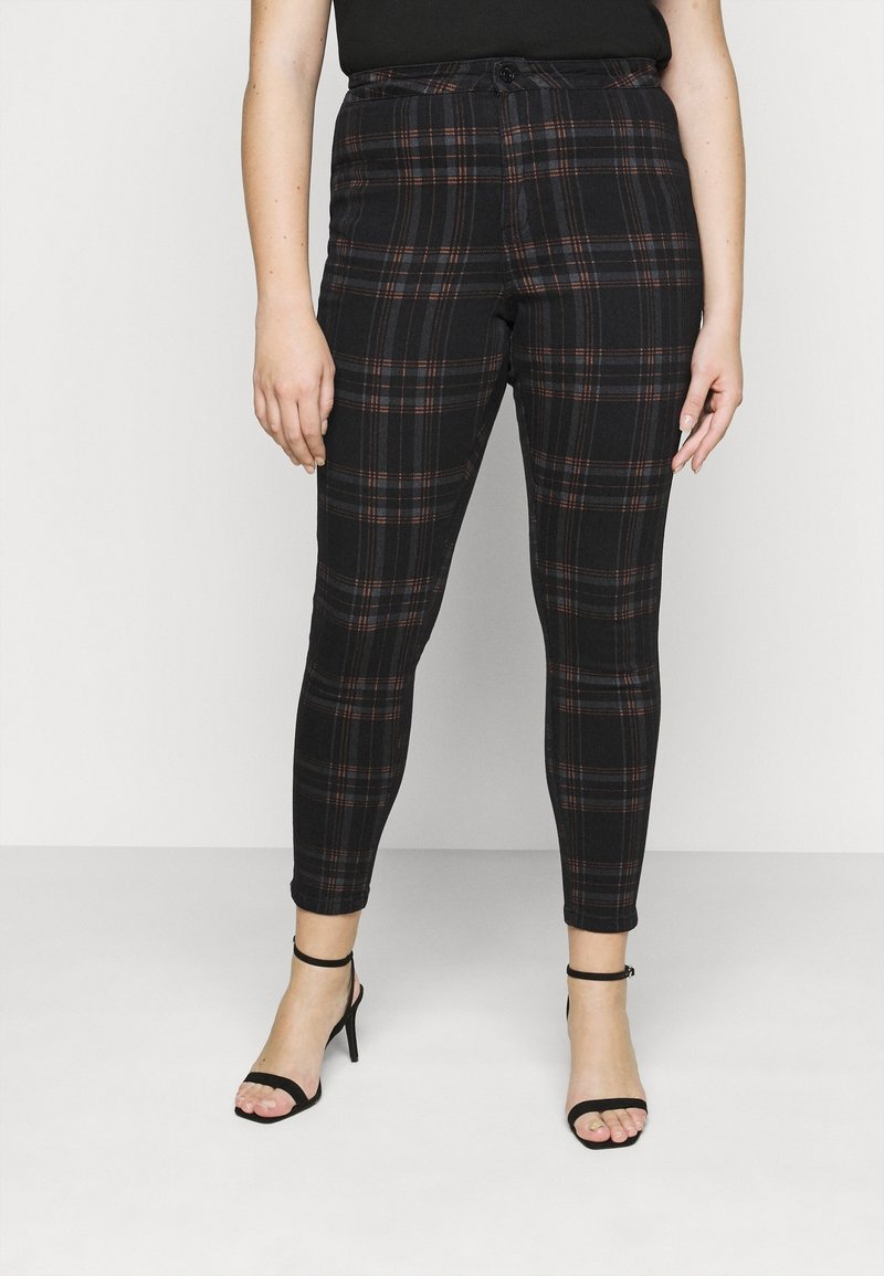 Mustat ruudulliset housut, joissa on punaisia ja sinisiä yksityiskohtia, kapeneva malli, korkea vyötärö ja nappikiinnitys, yhdistettynä mustiin remmi-korkokengiin.