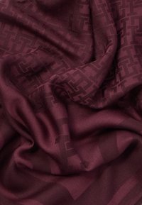 Dyb burgunderfarvet stof med subtile, mørkere geometriske mønstre og bløde folder, der skaber teksturerede, flydende overfladedetaljer.