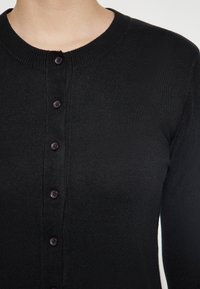 Musta neuletakki pyöreällä kaula-aukolla, jossa on nappikiinnitys edessä ja ribbitekstuuri. Nappit ovat tummat ja tasaisesti sijoiteltuja.