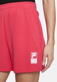 Los pantalones cortos de algodón rosa cuentan con una cinturilla elástica, bolsillos laterales y una impresión blanca del logo. La tela tiene una textura sutil y un diseño casual.