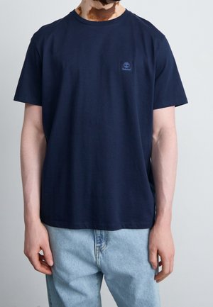 Mand iført en ensfarvet mørkeblå Timberland T-shirt og lyseblå jeans, stående mod en ensfarvet lys baggrund.